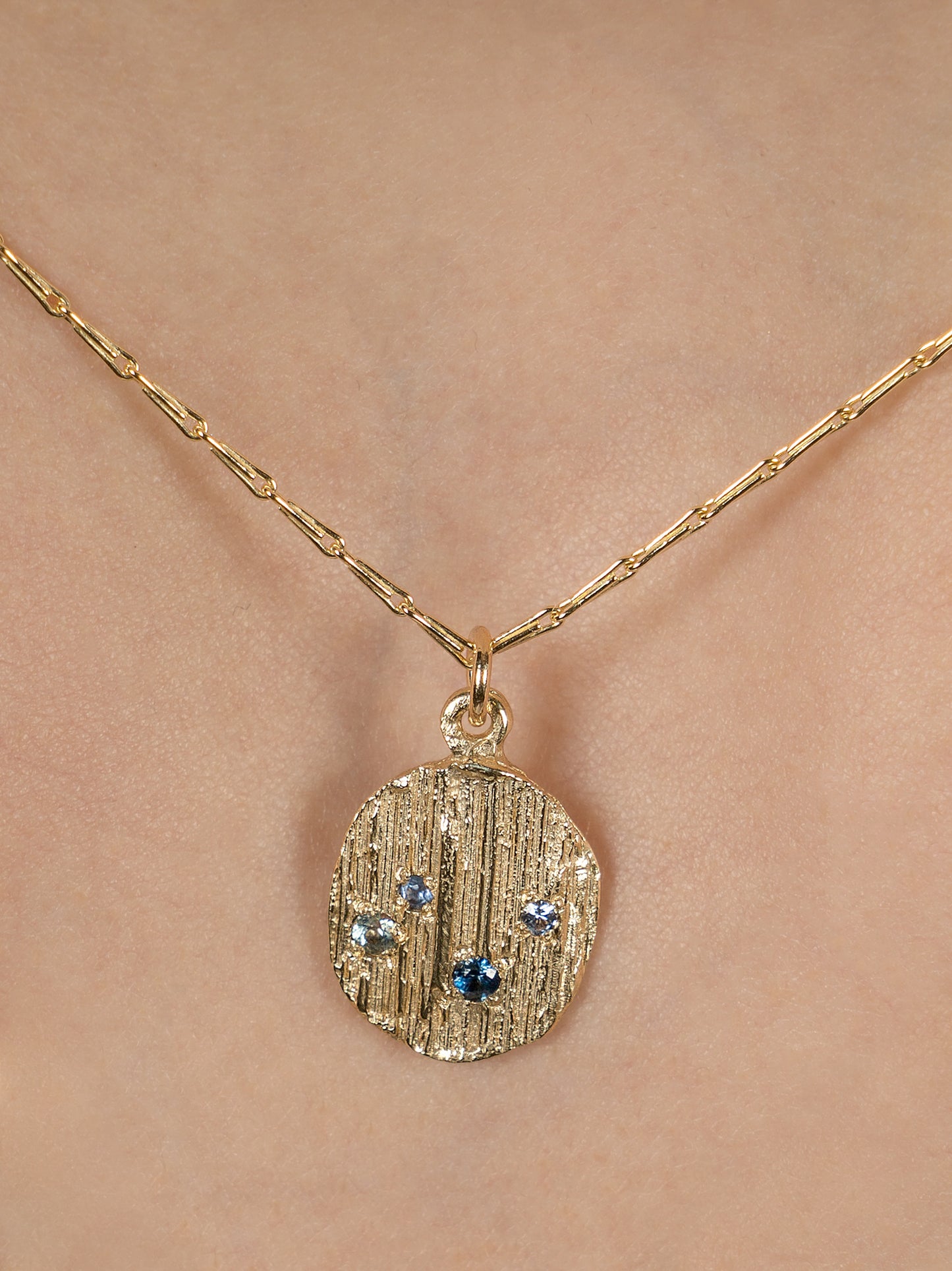 Picture of gold necklace with textured pendant set with 4 blue sapphires. Picture shows pendant being worn on model, close up image.