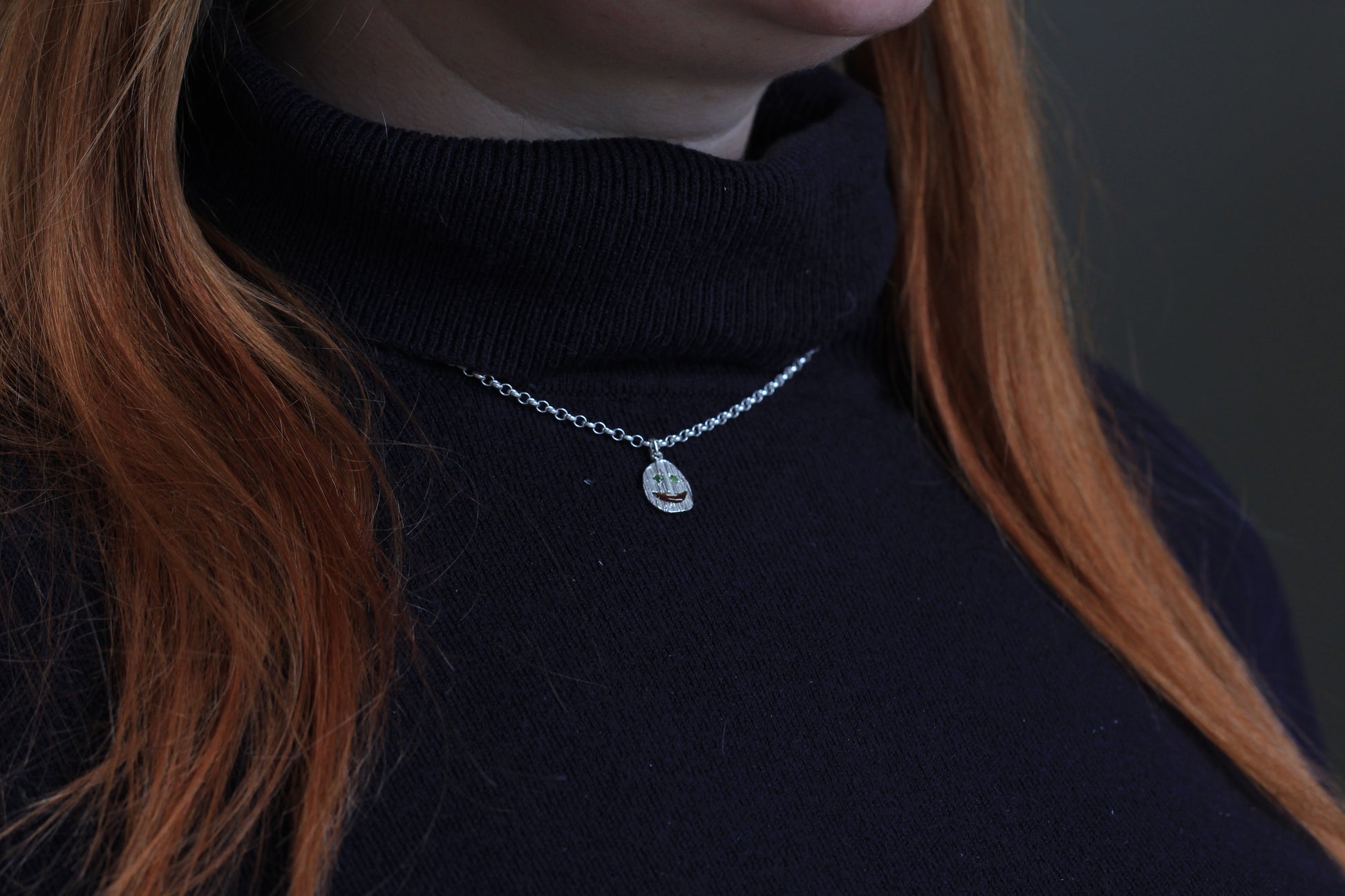 Picture of a model wearing a silver necklace with a smiley pendant. Eyes ahve green gemstones and mouth is enamelled.