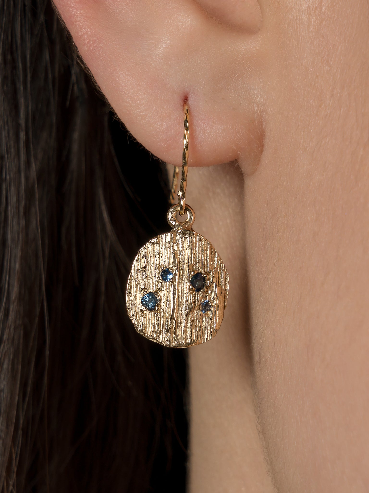 Close up picture on models ear of blue sapphire bark earrings in gold. 