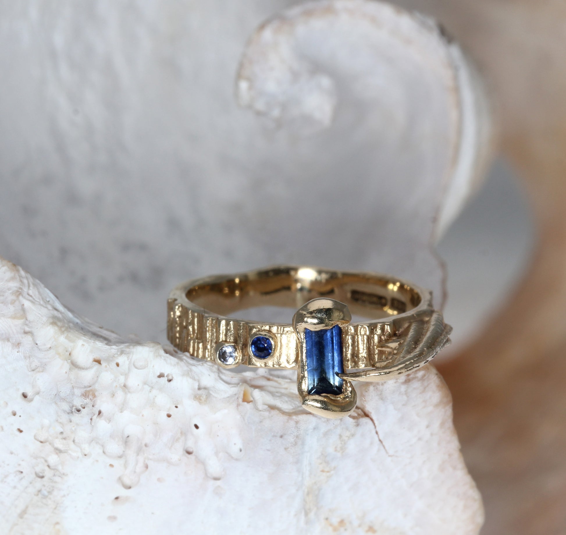 Gold textured contemporary engagement ring with blue sapphire gemstones on a shell background 