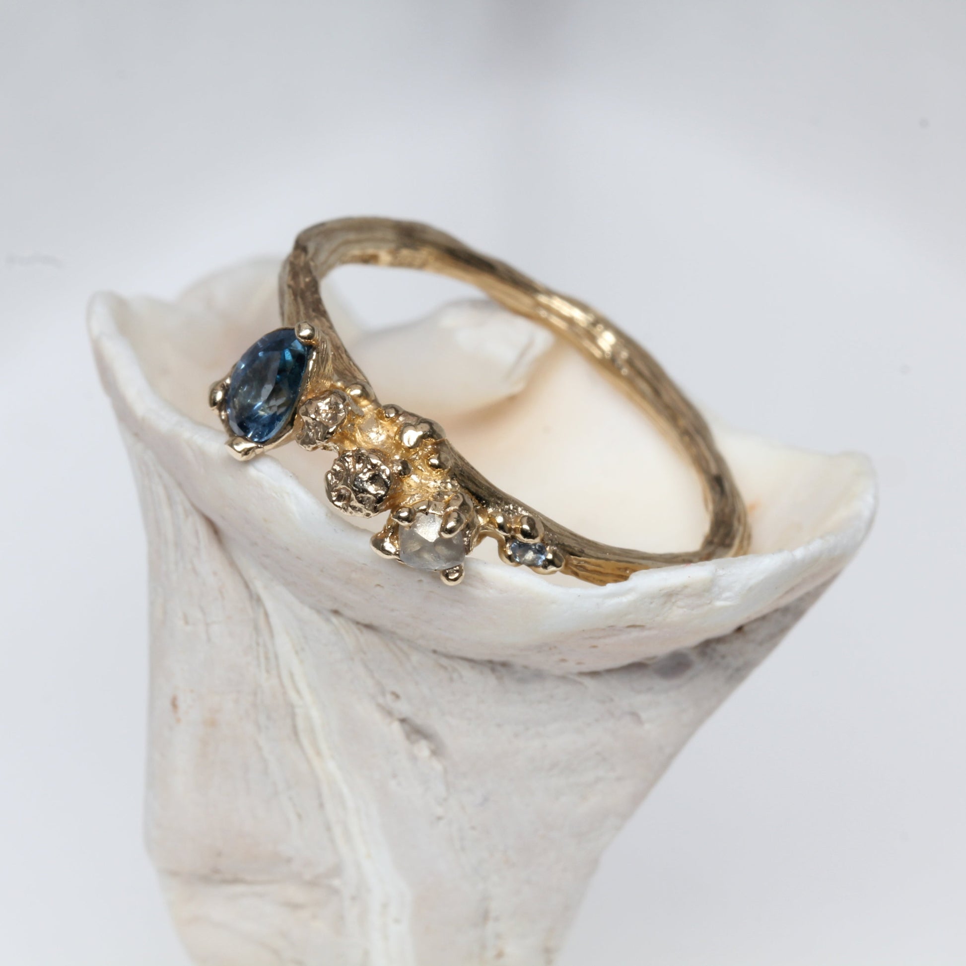 Gold organic engagement ring with a blue gemstone and raw diamond on a white shell against a light gray background