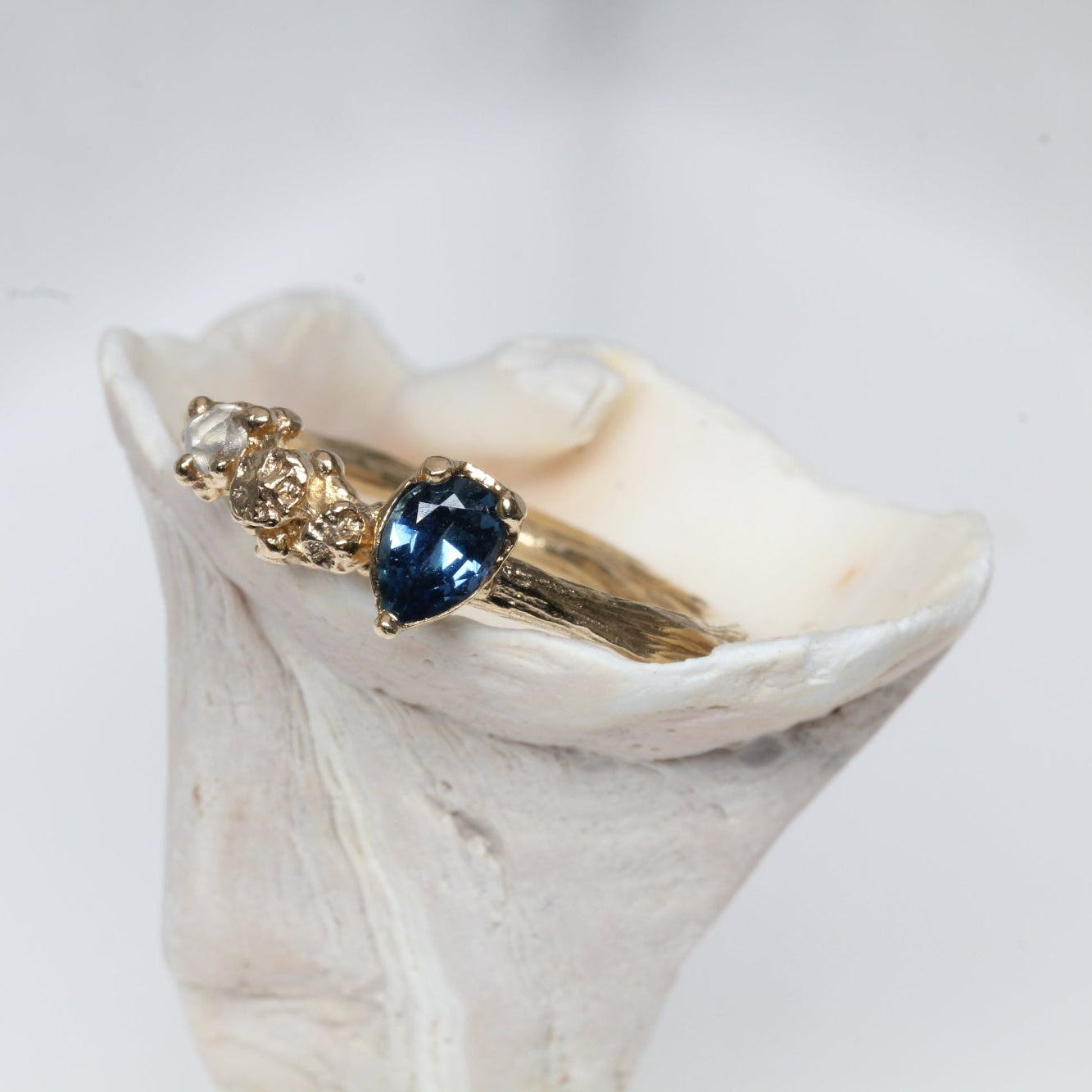 Gold engagement ring with a blue sapphire gemstone and raw diamond  on a white shell against a light gray background