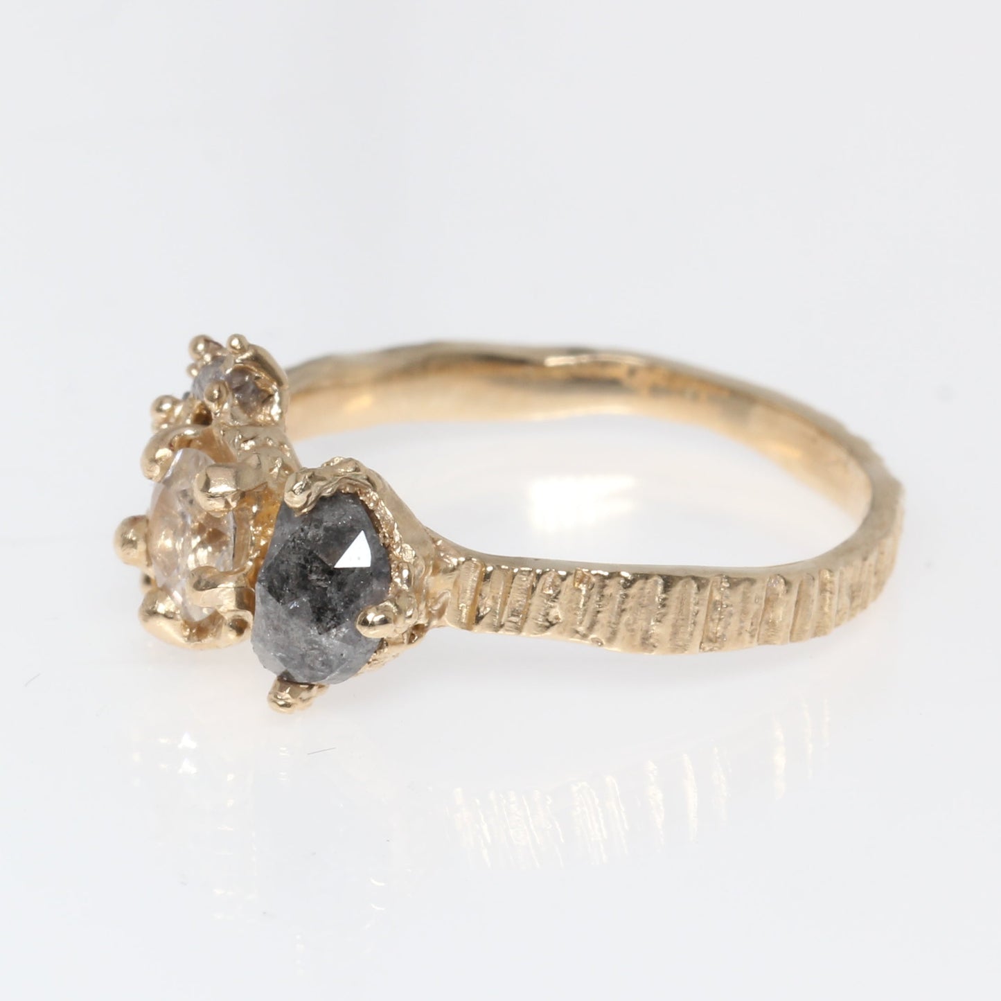 A yellow gold ring featuring a central round white sapphire, side stone is dark gray diamond  set on a textured band, shown on a plain white background.