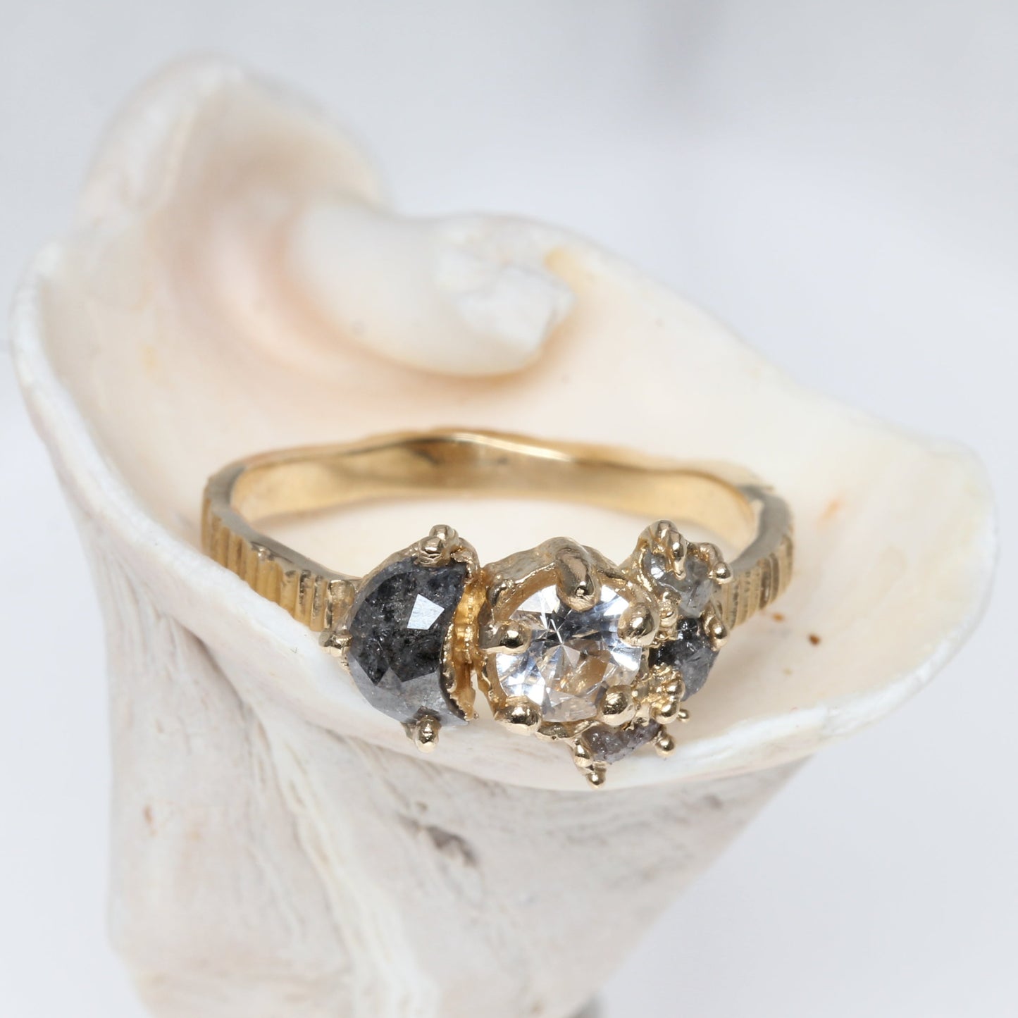 A gold three-stone ring with a clear center stone and dark diamond side stones, displayed on a white clam shell against a white background.