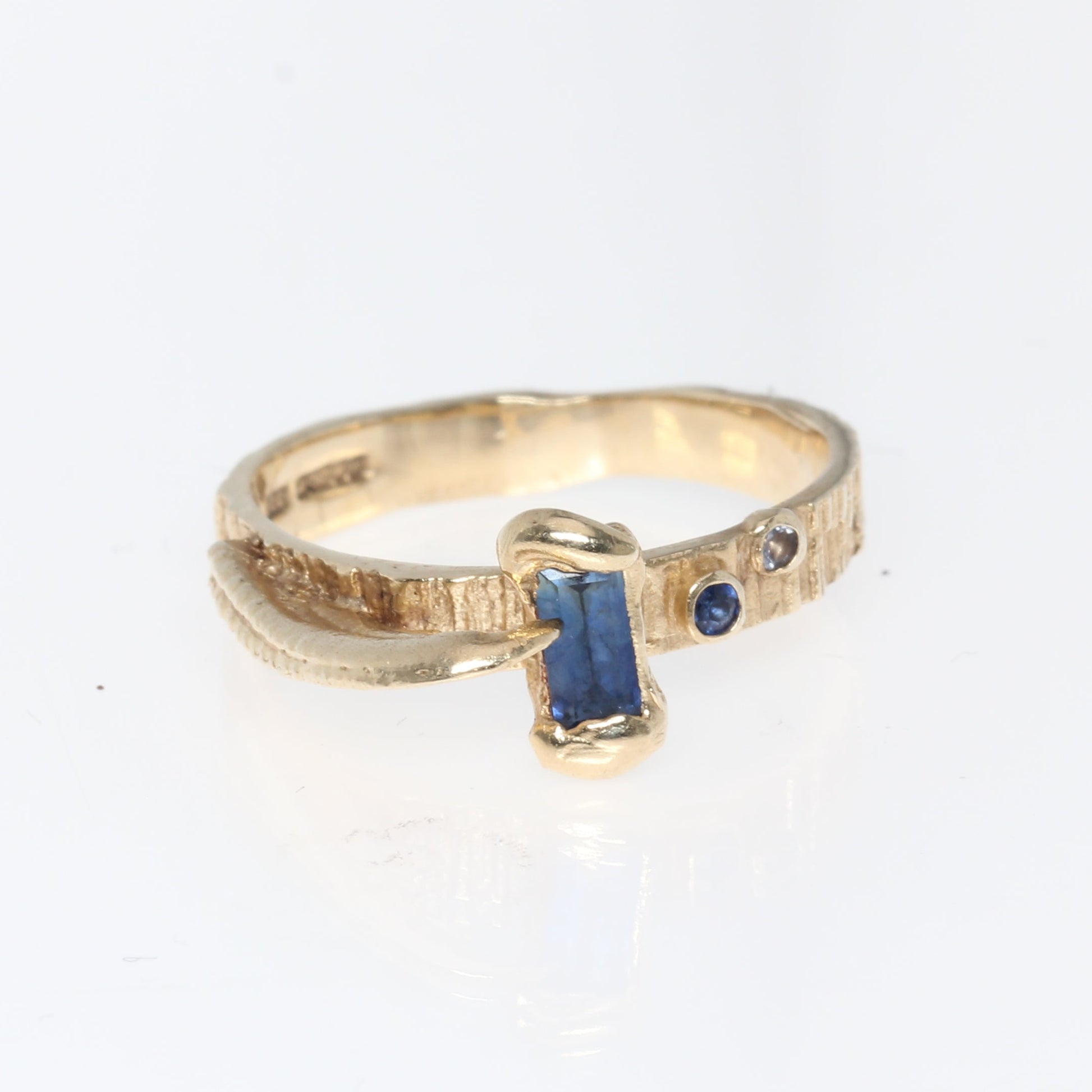 A gold ring with a central rectangular blue sapphire and small side accents, photographed on a plain white background.
