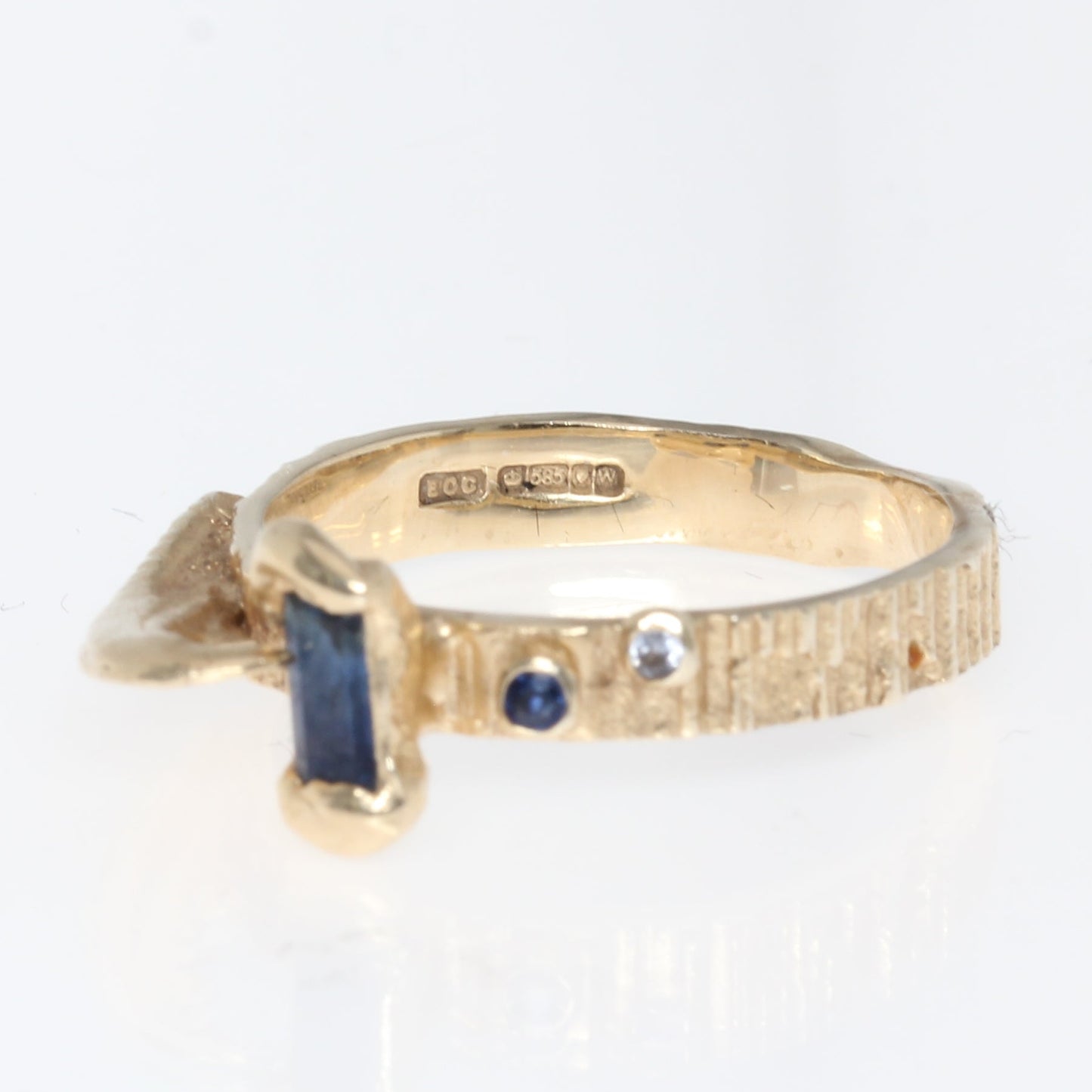 A yellow gold ring with a textured band, featuring a rectangular blue gemstone on one side and small round blue and clear stones on the other, set against a plain white background. Hallmark shown here. 