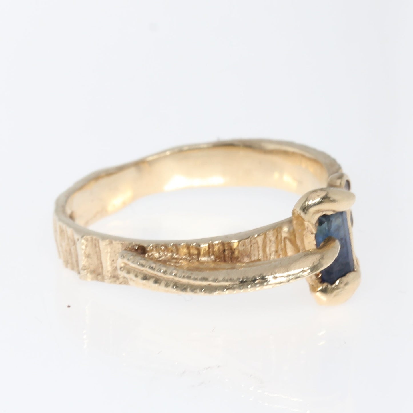 A gold ring with a textured band and a small dark blue rectangular gemstone, shown on a plain white background.