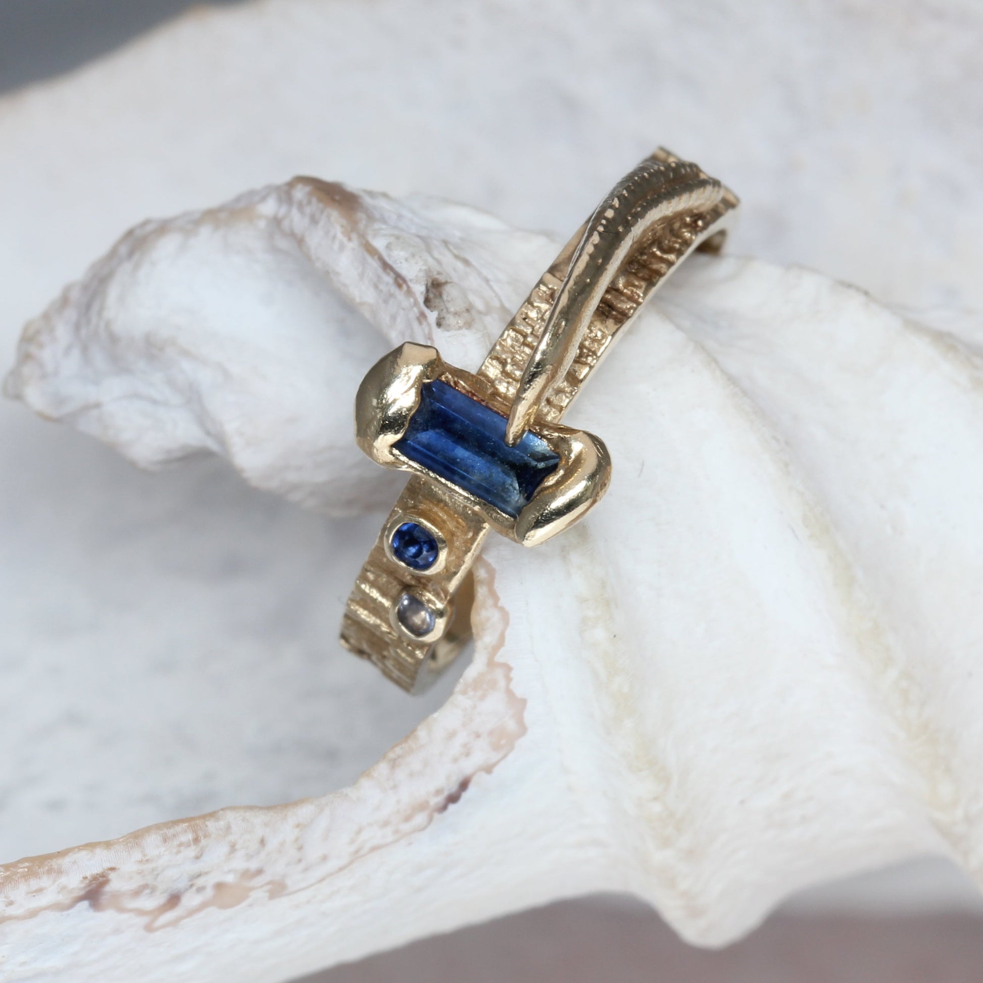 A gold ring with a central rectangular blue gemstone and small accent stones, displayed on a textured white shell.
