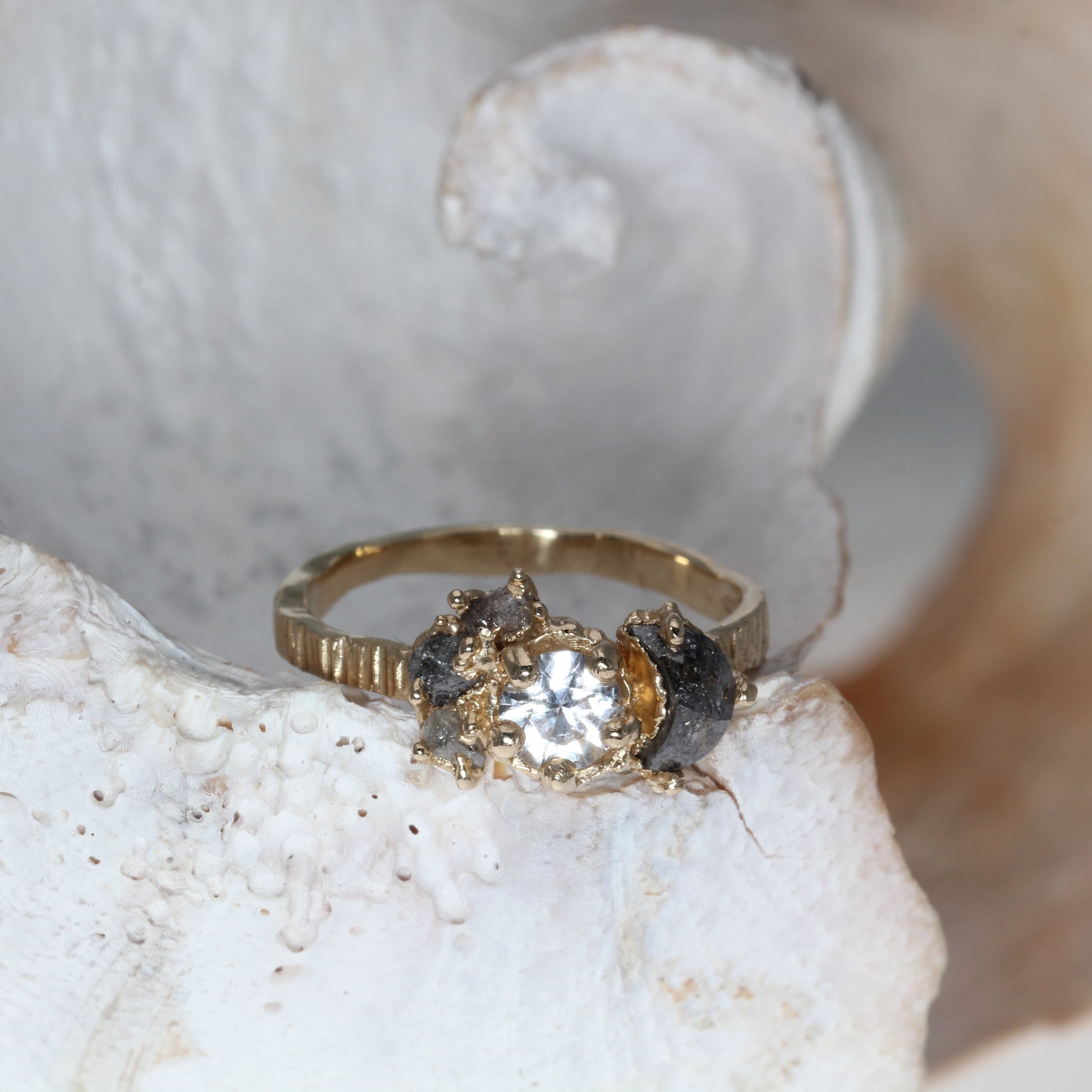 A yellow gold ring with a central clear round gemstone and dark side stones, displayed on a textured white seashell.