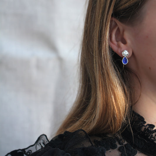 Silver earrings in the shape of crab shells with blue enamel drop that looks like a sapphire gemstone shown here on a model. 