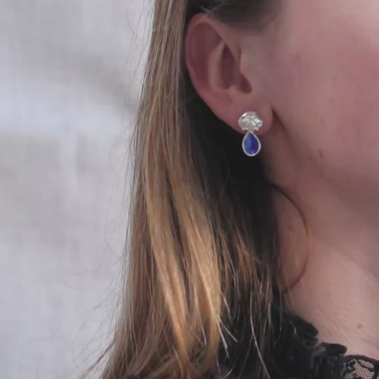 Silver earrings in the shape of crab shells with blue enamel drop that looks like a blue sapphire gemstone shown here on a model. 